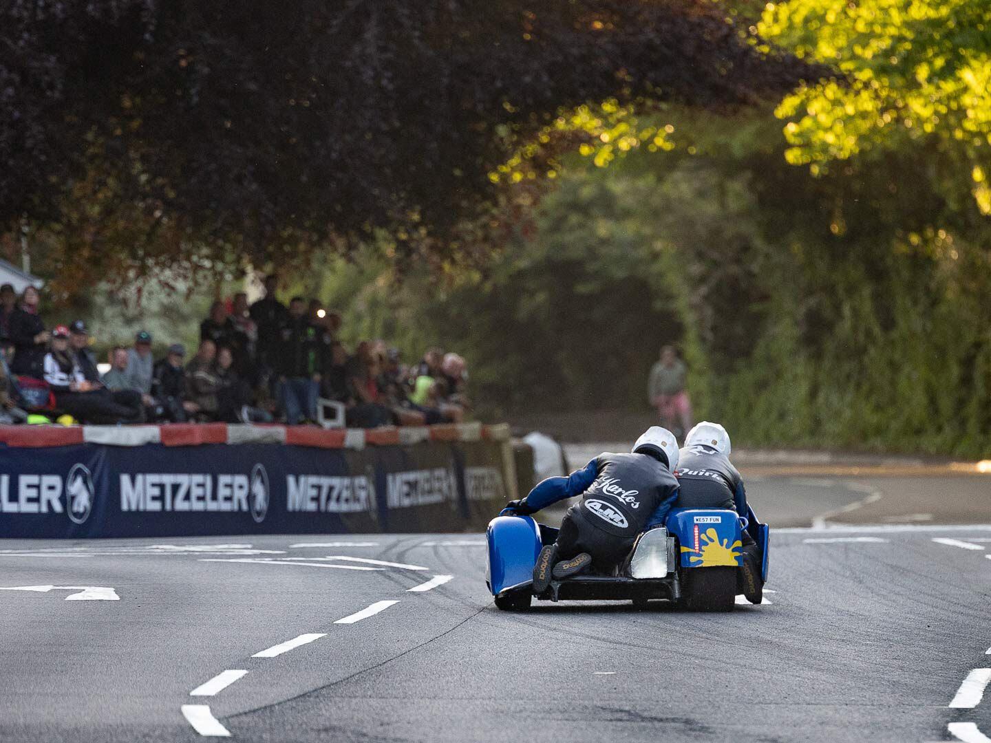 Low to the ground and close to the crowd, monkey Karl Schofield moves from side to side to balance the sidecar as pilot Dave Quirk navigates the Suzuki power machine past Braddan Church.